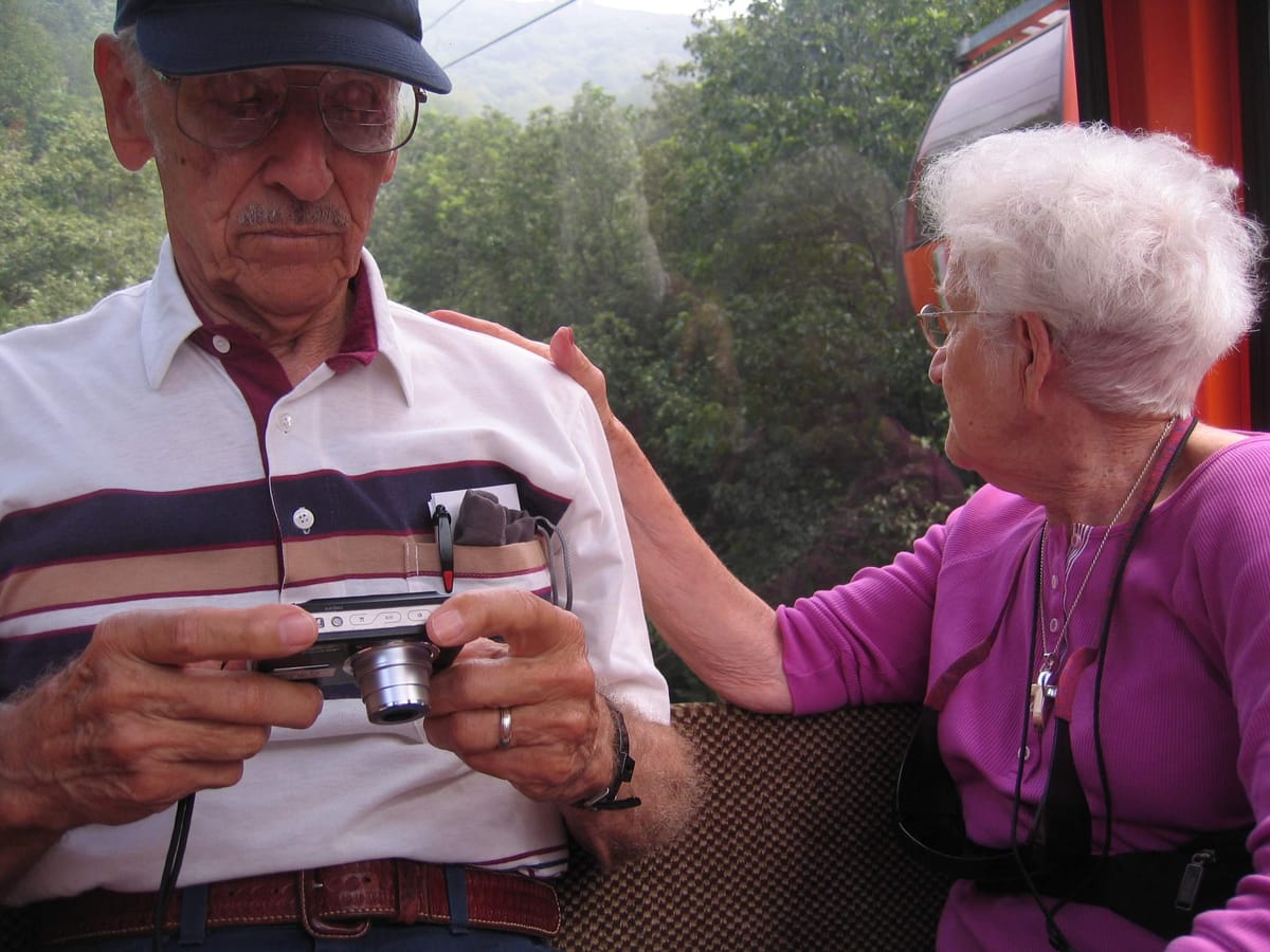 Sitting in a gondola, Granddad reviews a photo on his digital camera while Grandmom rests a hand on his shoulder and looks out over the trees
